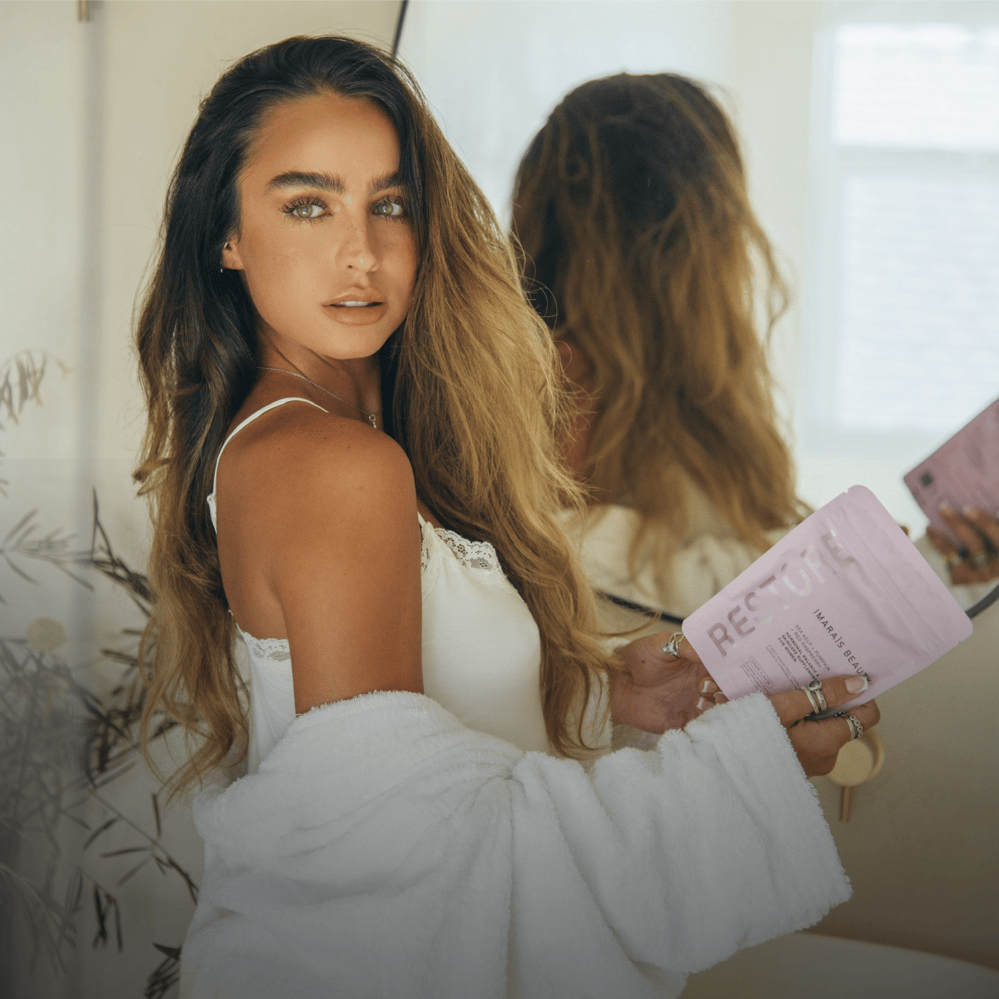 Model Sommer Ray posing indoors with long, wavy hair, wearing a white tank top and robe, holding an Imarais Beauty Restore gummy supplement pouch.