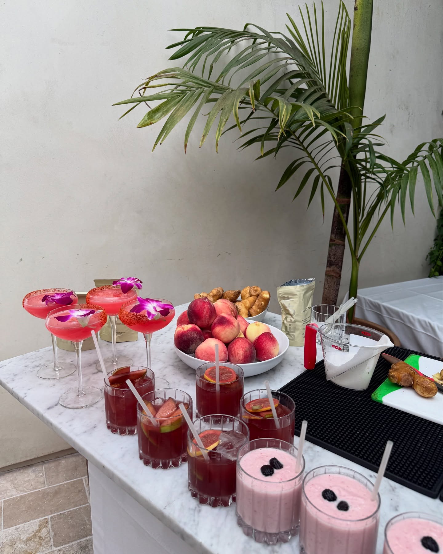 Outdoor setup featuring colorful wellness drinks, peaches, fresh ginger, and IMARAÏS BEAUTY pouches on a marble table, surrounded by tropical greenery.