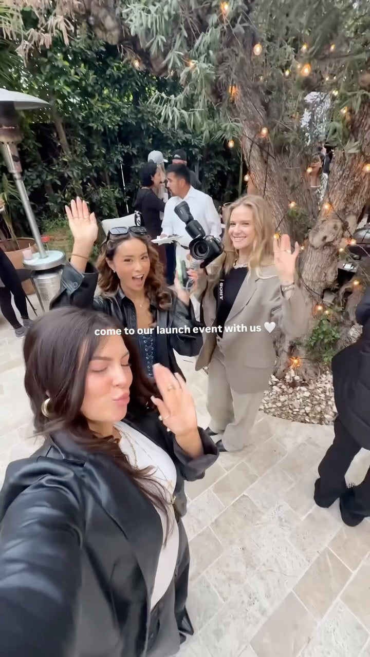"Group of women smiling and waving at an outdoor IMARAÏS BEAUTY launch event, surrounded by twinkle lights, trees, and other guests, with text overlay reading 'come to our launch event with us 🤍